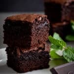 Fresh mint brownies stacked on a white plate with mint and chocolate near.