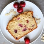Cherry almond bread on a white plate with fresh cherries and sliced almonds on the side.