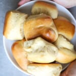 Woman holding a white bowl with dinner rolls in it.