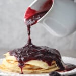 Pouring blackberry syrup over a stack of pancakes on a white plate.