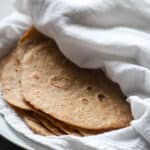 Stack of einkorn tortillas wrapped in a white dish towel on a white plate.