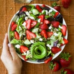 Woman holding a bowl of green salad with strawberries and feta with an antique fork on the side.