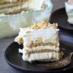 Slice of banana icebox cake on a black plate with a glass pan behind it.