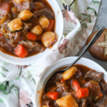 Pressure cooker beef stew in white bowls with a napkin under them and antique spoons on the side.