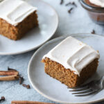 Slices of gingerbread cake with cream cheese icing on small plates with fresh cinnamon on the side.