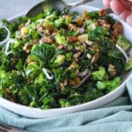 Kale and broccoli salad in a white bowl with a woman holding a spoon next to it.