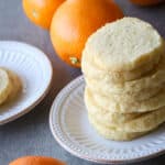 Orange shortbread cookies stacked on a white plate with navel oranges on the side.