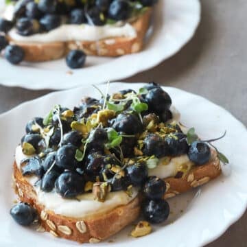 Ricotta and blueberry toast on a white plate topped with pistachios.