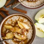 Apple cinnamon porridge in ceramic bowls with cinnamon sticks and sliced apples on the side.