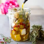 Woman pouring olive oil into a mason jar with feta and other herbs in it with flowers in the background.