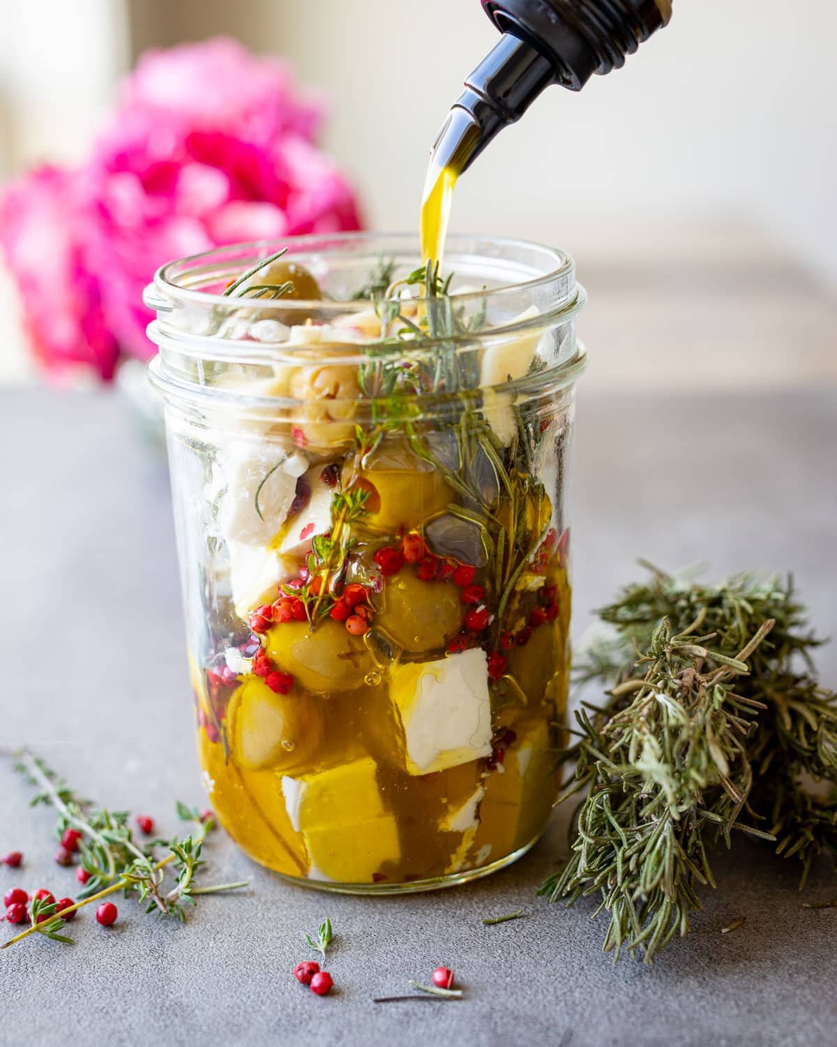 Woman pouring olive oil into a mason jar with feta and other herbs in it with flowers in the background.