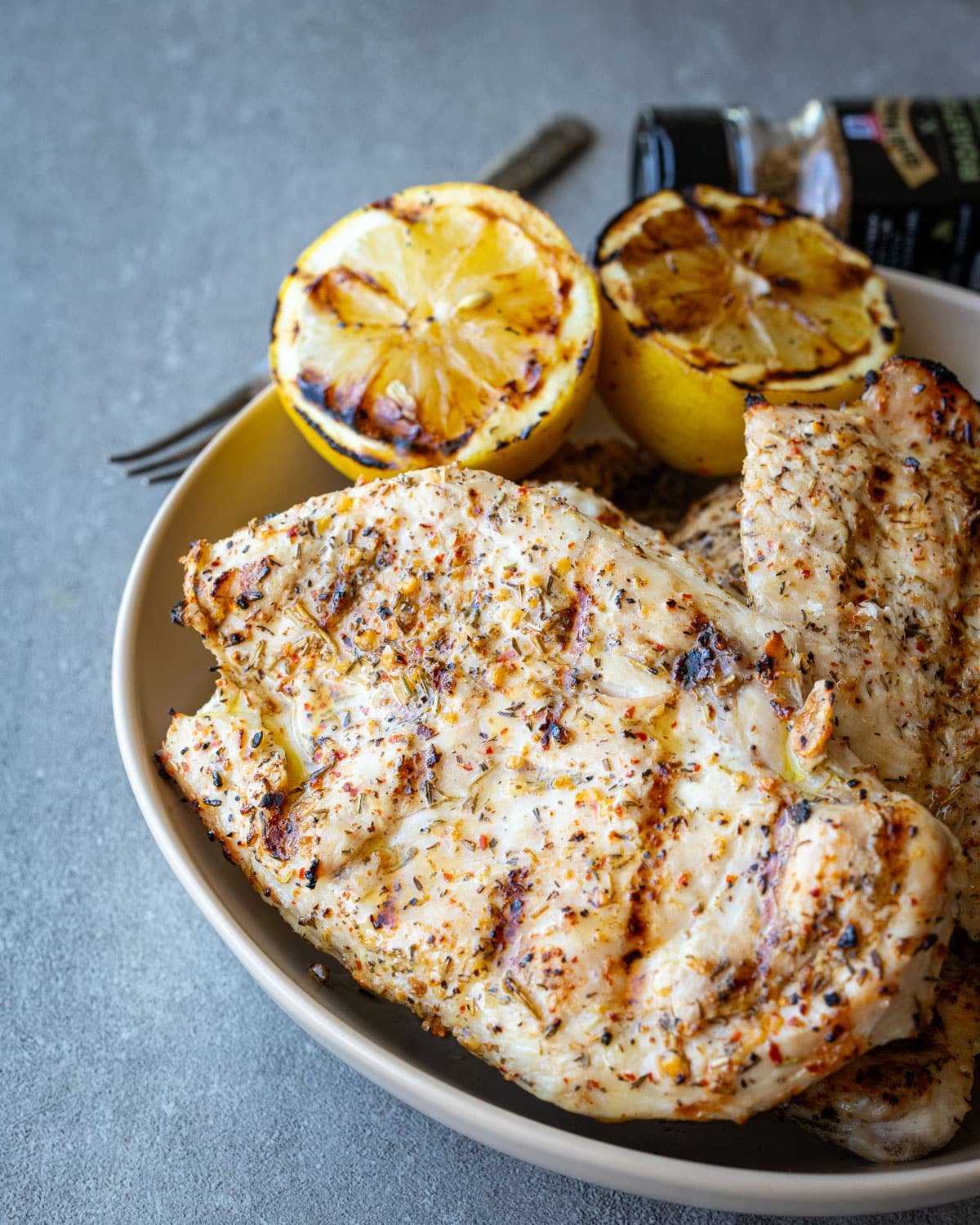 Grilled Italian herb chicken cutlets stacked in a ceramic bowl with grilled lemon halves on the side. 