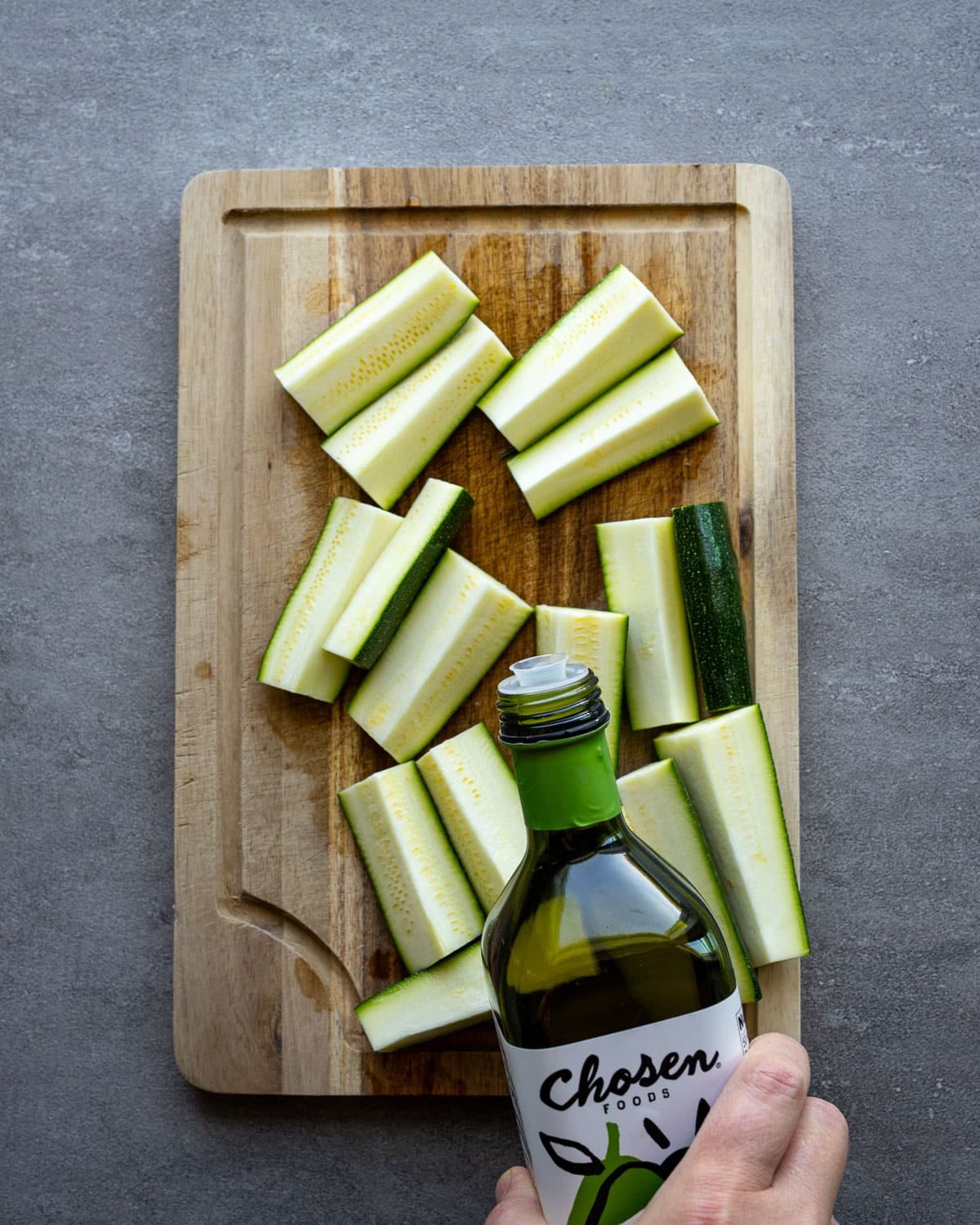 Woman drizzling avocado oil over zucchini on a wooden cutting board.
