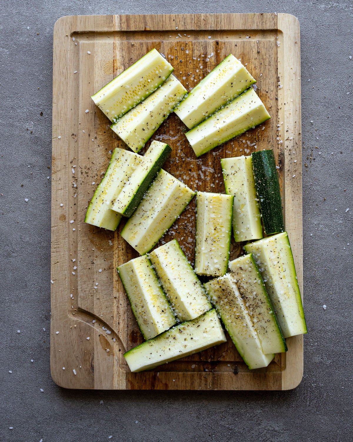 Zucchini sprinkled with salt and pepper on a wooden cutting board.