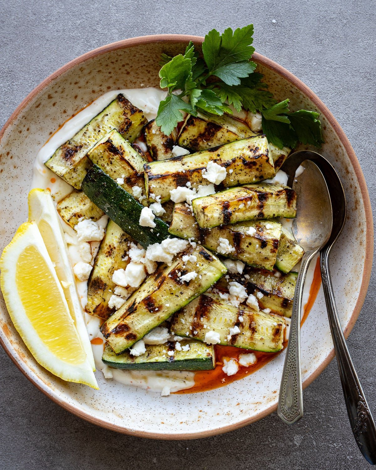Grilled zucchini in a ceramic bowl with lemon wedges and fresh parsley on the side.