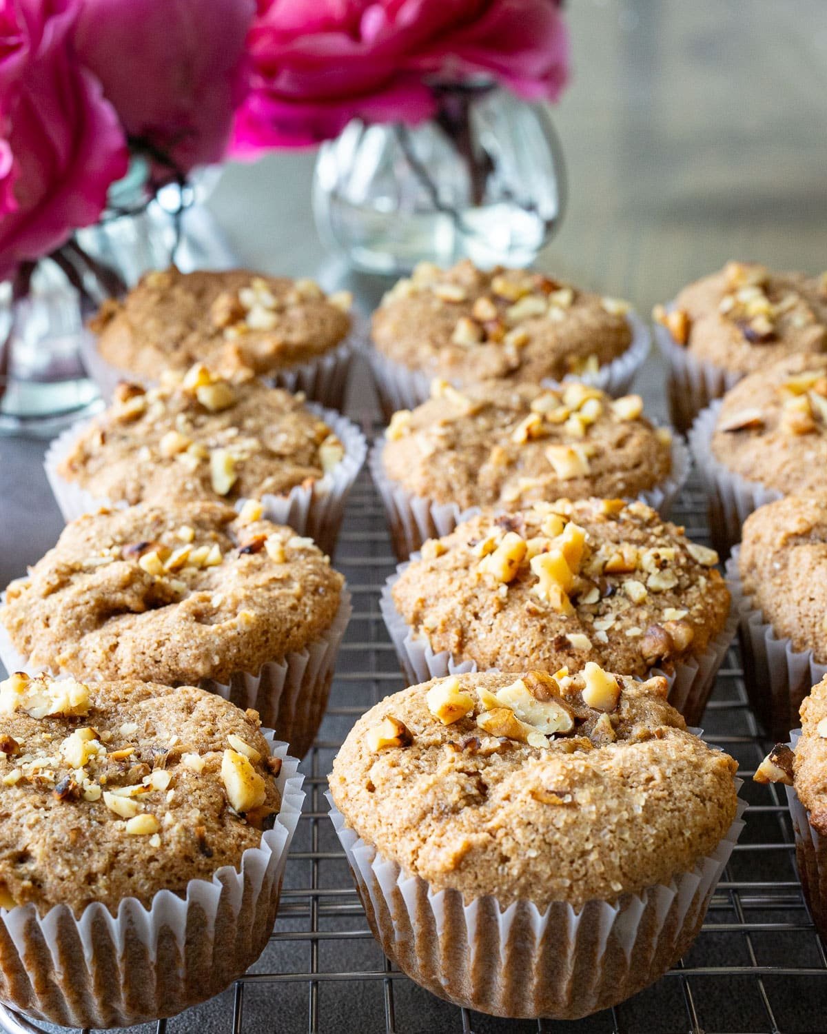 Sprouted wheat flour muffins topped with chopped walnuts on a cooling rack with pink flowers in the background. 
