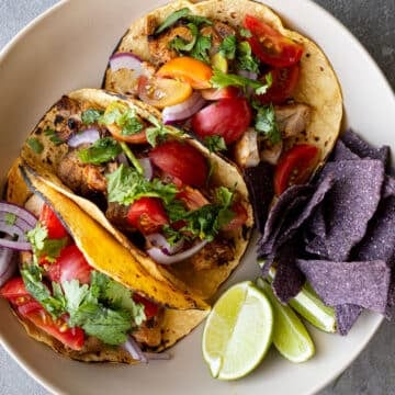 Grilled chicken tacos on a white plate with blue corn chips on the side.