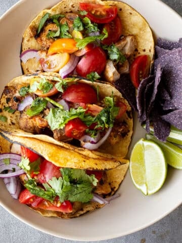 Grilled chicken tacos on a white plate with blue corn chips on the side.