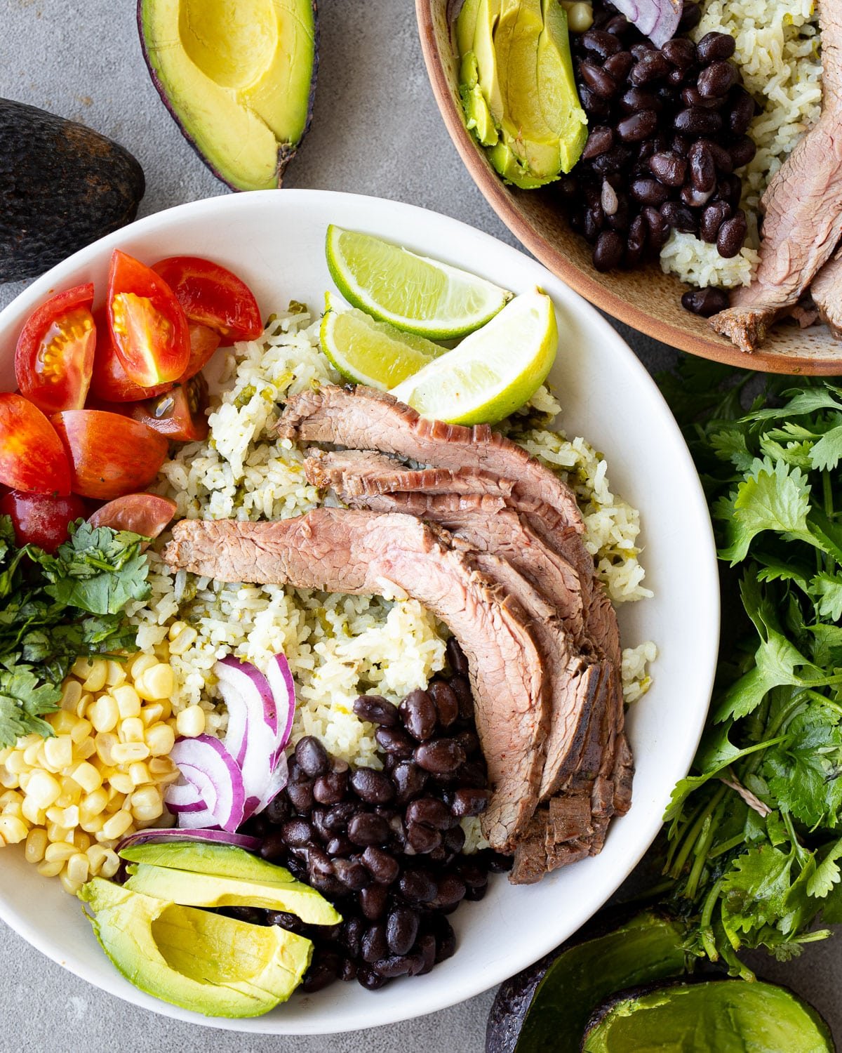 Carne asada bowls in ceramic bowls with avocados, lime, and cilantro on the side.